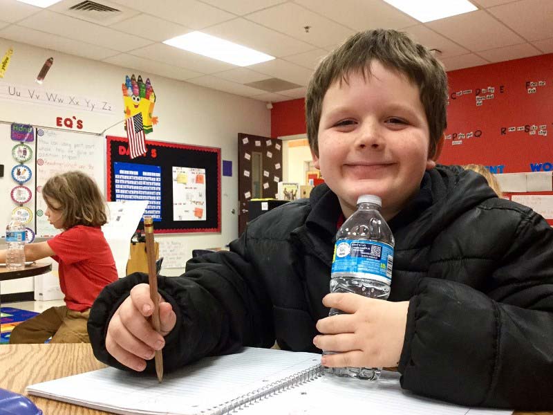 Smiling male elementary student with a bottled water touching his chin