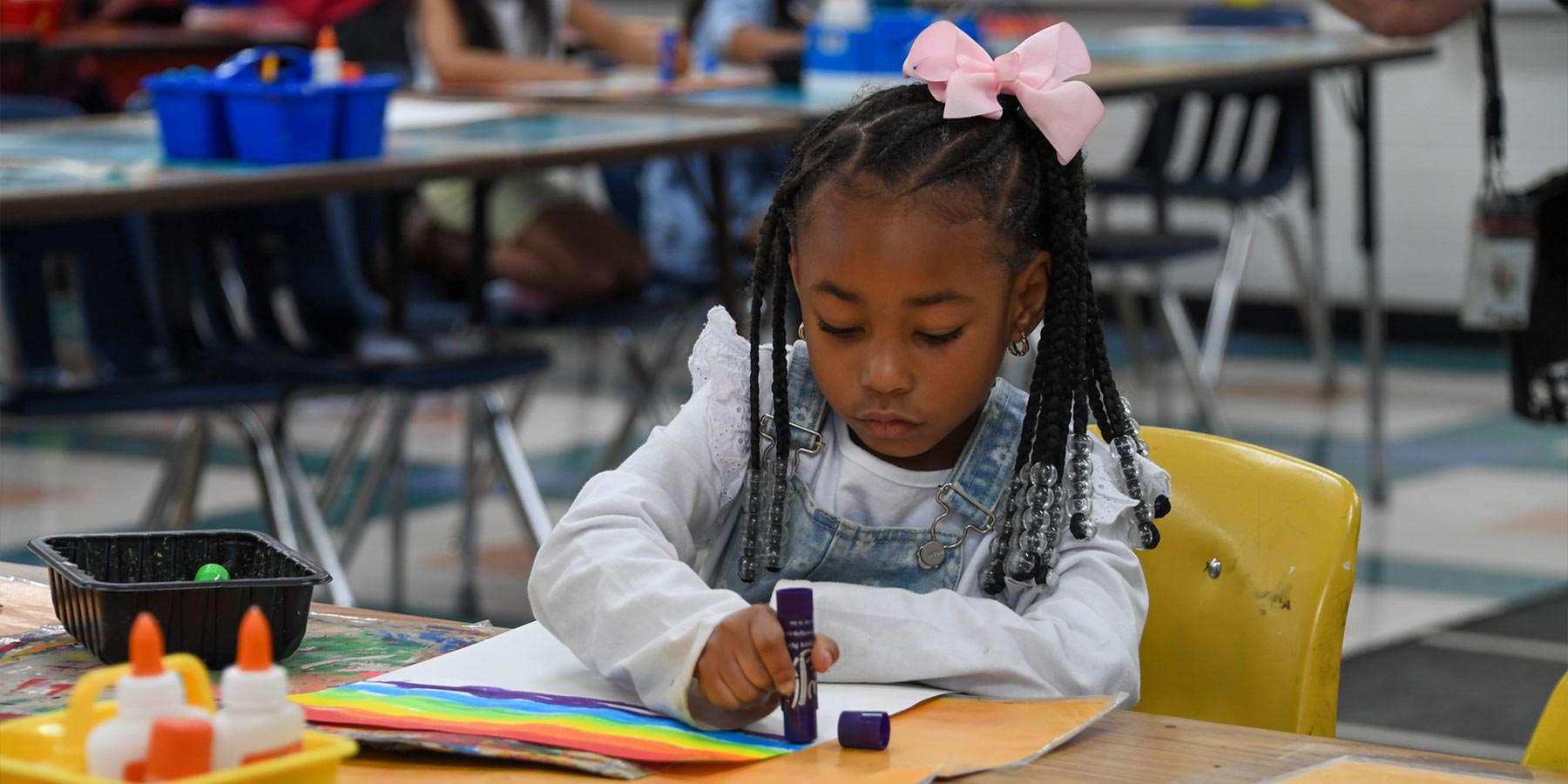 elementary girl using glue stick with artwork at a classroom table