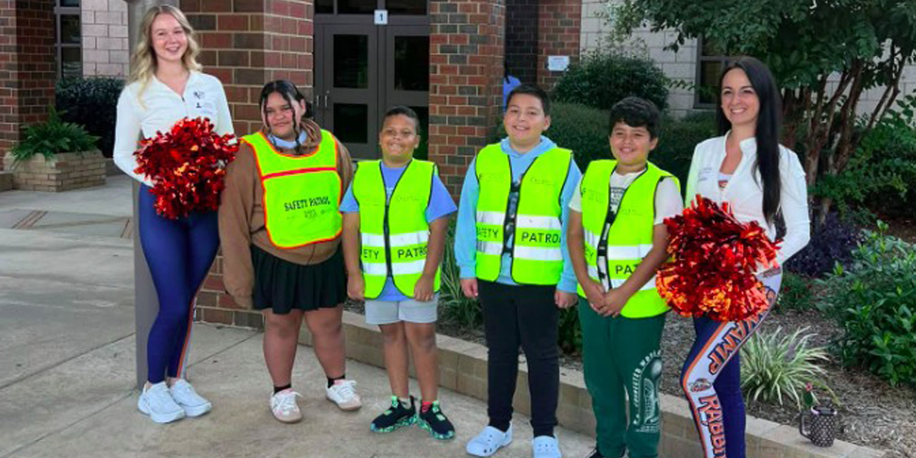 4 safety patrol students between two teachers holding red pom poms